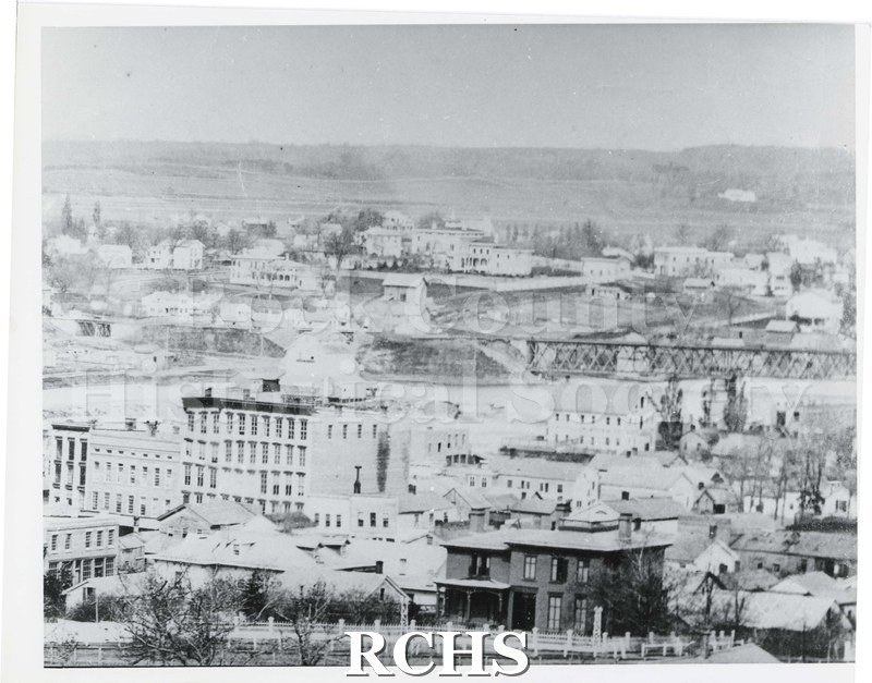 Black and white photograph of downtown Janesville. Background buildings are not in focus. There is a two story house in front with fence in front of it.