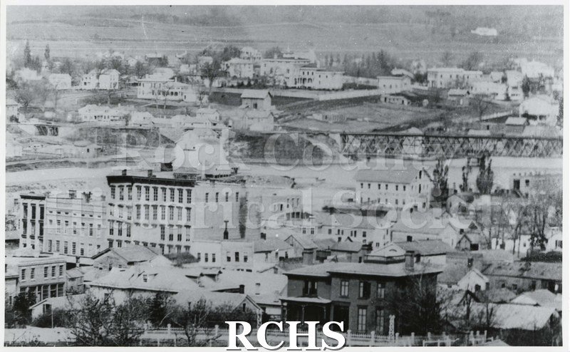 Black and white photograph of downtown Janesville, WI. Background buildings are not in focus. There is a two story house in front with fence in front of it.