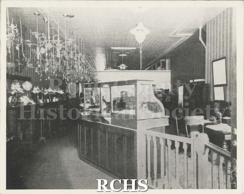 Black and white photograph of Gas Light Company-Now Wisconsin Power and Light. Shows inside of a building with hanging light fixtures and lamps on tables.