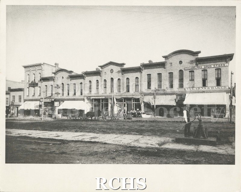 Black and white photograph of 109-123 West Milwaukee Street featuring 'Old Town Pump'". On top of building to the right is a sign: "Philadelphia Drug Store".