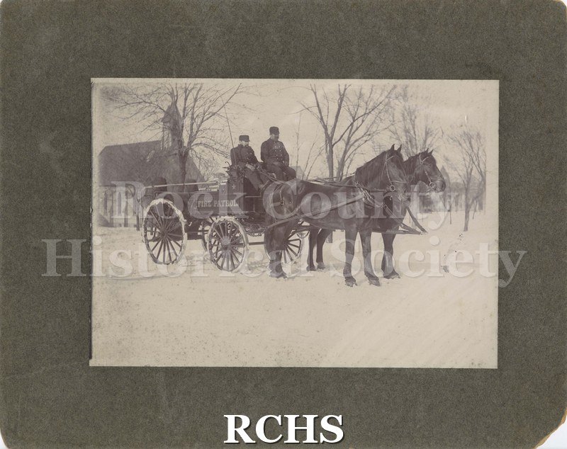Black and white photograph of Janesville Fire Patrol. Two men are in a wagon pulled by two horses. On side of wagon is "Fire Patrol". Church is in the background.