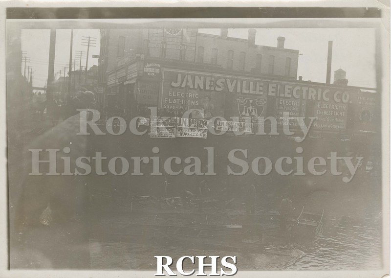 Black and white photograph of the Janesville Electric Co. sign. Photo is quite blurry but large building is behind sign.