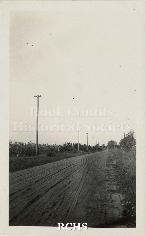 Black and white photograph of rural electrification. Shows a rural road with electrical poles. Road is rutted and there is a corn field on the left behind the poles.