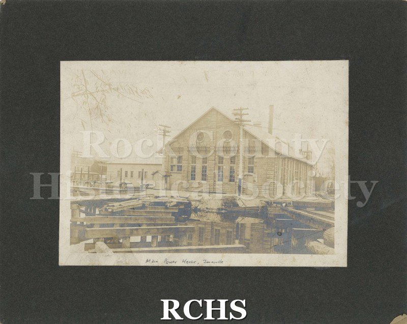 Black and white photograph of the Main Power House, Janesville. Shows the building with power poles in front and smokestack in back and wooden planks in front.
