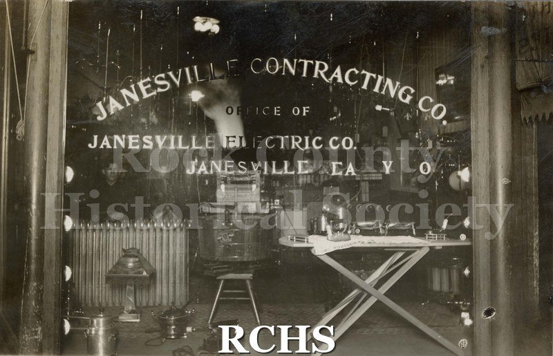 Black and white photograph of Janesville Electric Co. Shows the interior of a building with sign on window: Janesville Contracting Co.
