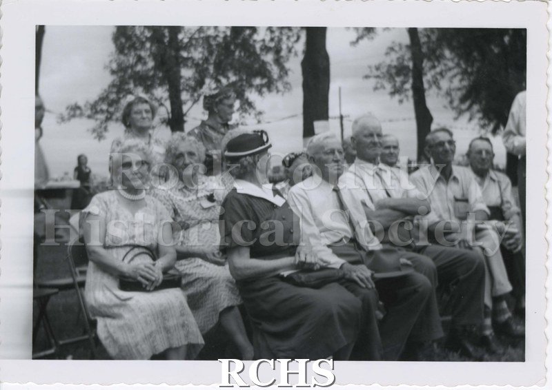 Black and white photograph of West Center School Reunion. Older men and women sit and stand facing the camera, except for one woman turned away. All are dressed up.