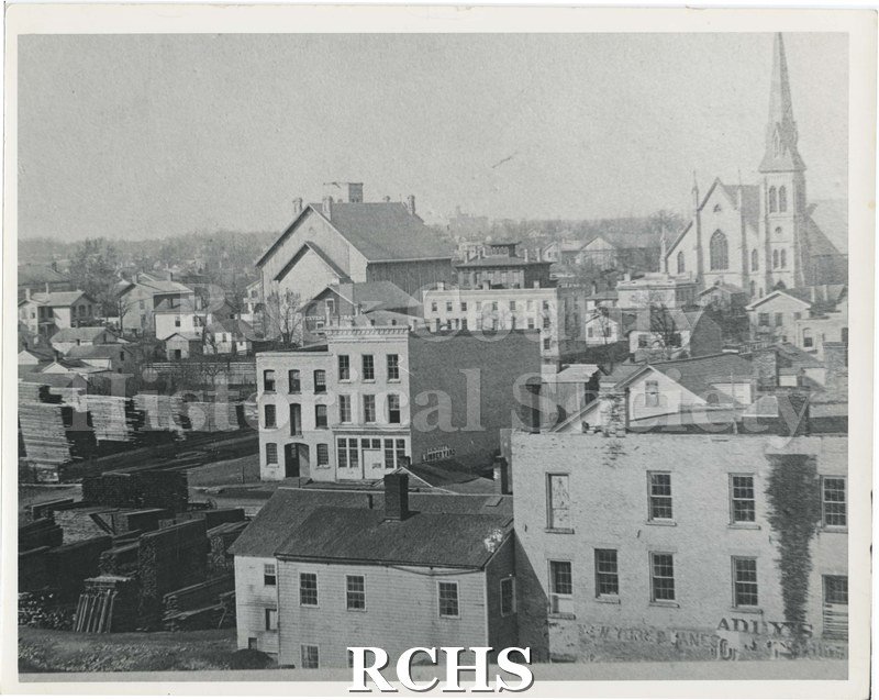 Black and white photograph of Janesville Downtown. Churches, lumberyard, building with a sign reading, "Addy's New York [illegible].".