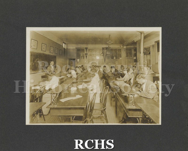 Black and white photograph of Janesville Business College 1911. It shows men and women students at work at desks with teacher standing in front of a blackboard.