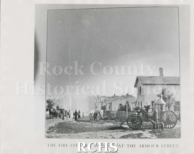 Black and white photograph of the Fire Steamer at work at the Armour Street Well. Shows houses on right. Group of men visible to the left, likely firefighters.