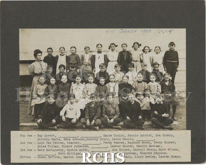 Black and white photograph of fourth grade 1907. Shows a fourth grade teacher and her class standing outside a building. Circa 1907.