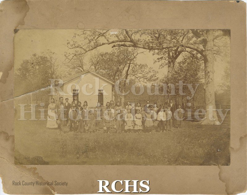 Black and white photograph of Merrifield School, class photo, 1874. Students are standing in front of a white, one-room schoolhouse.