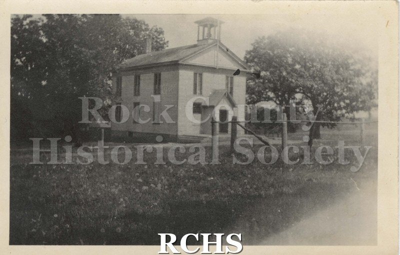 Black and white photograph of Shopiere grade school, two floors with a cupola. Fence in front, trees in background.