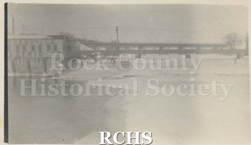 Black and white photograph of the upper bridge and dam, Janesville, with old Power House. Janesville Electric Company is written on the building.