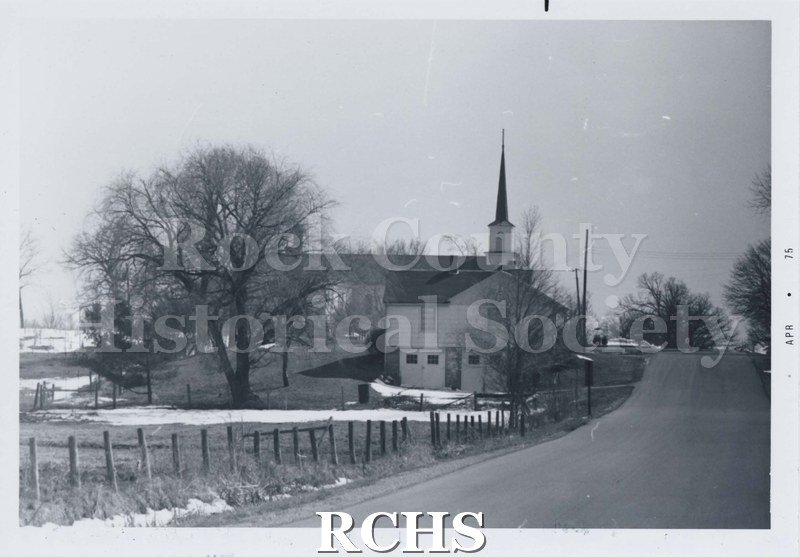 Black and white photograph of the Newark Cheese Factory. Shows a street and field. A church with a steeple in background, large building in foreground.