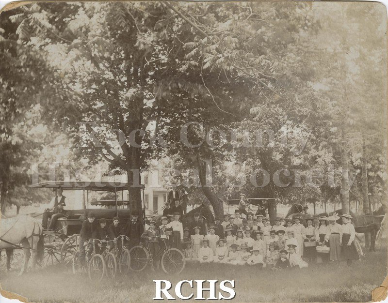 Black and white photograph of Evansville graded school, around 1900 or earlier. The students stand outdoors under trees, house in the background.