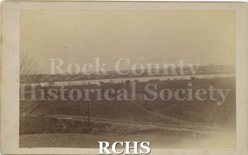 Sepia aerial photo of Janesville over looking the Rock River (?). The edges are slightly faded, but the scene is of fields (farming?) that lead up to the river.