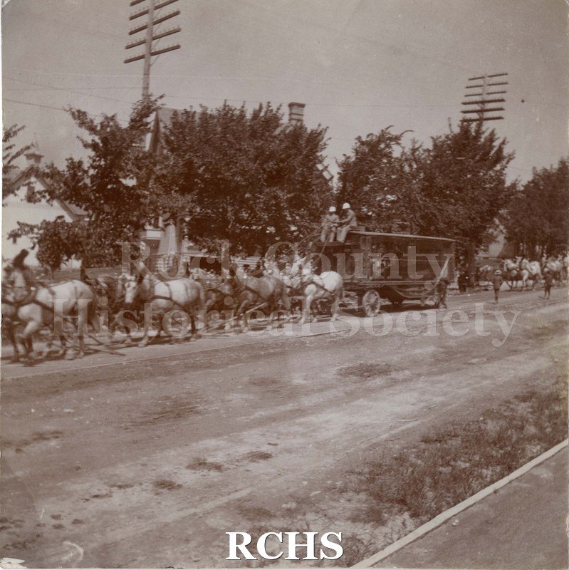 Black and white photograph of a Circus float, maybe for animals, drawn by eight horses. Two men ride on top of the cart or float.