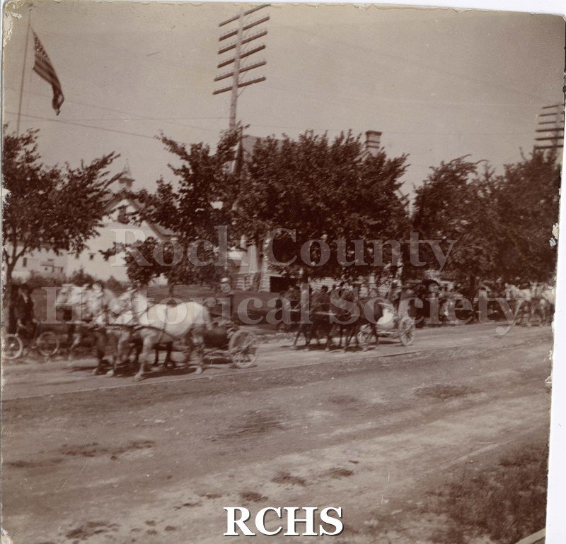 Black and white photograph of groups of four horses drawing chariots in a circus parade while people watch from the side of the road.