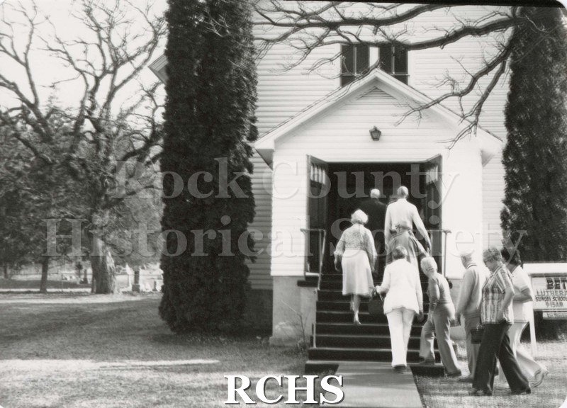 Black and white photograph of men and women entering a white, wooden building. Possibly a church?