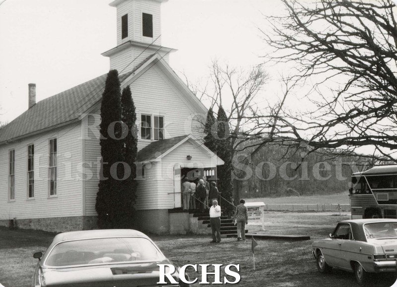 Black and white photograph of men and women entering a white, wooden church with a steeple. Cars parked in front.