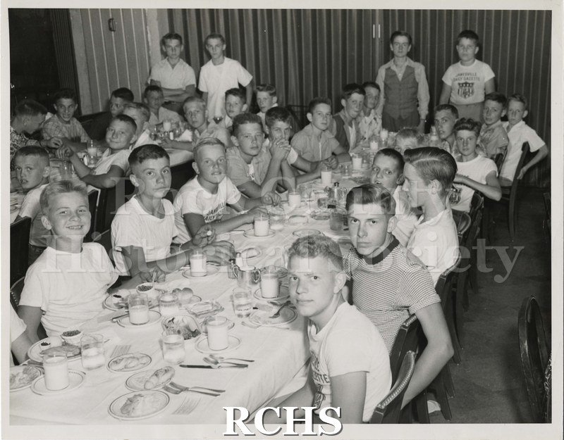 Black and white photograph of Carliss Shuler and local boys eating dinner at a Soap Box Derby function. Many have shirts reading "Janesville Daily Gazette.".