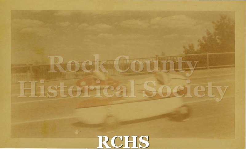 Tinted photo of three soap box cars racing over Monterey Bridge in Janesville. Soap Box Derby race, 1948. Carliss B. Shuler is in the left car.