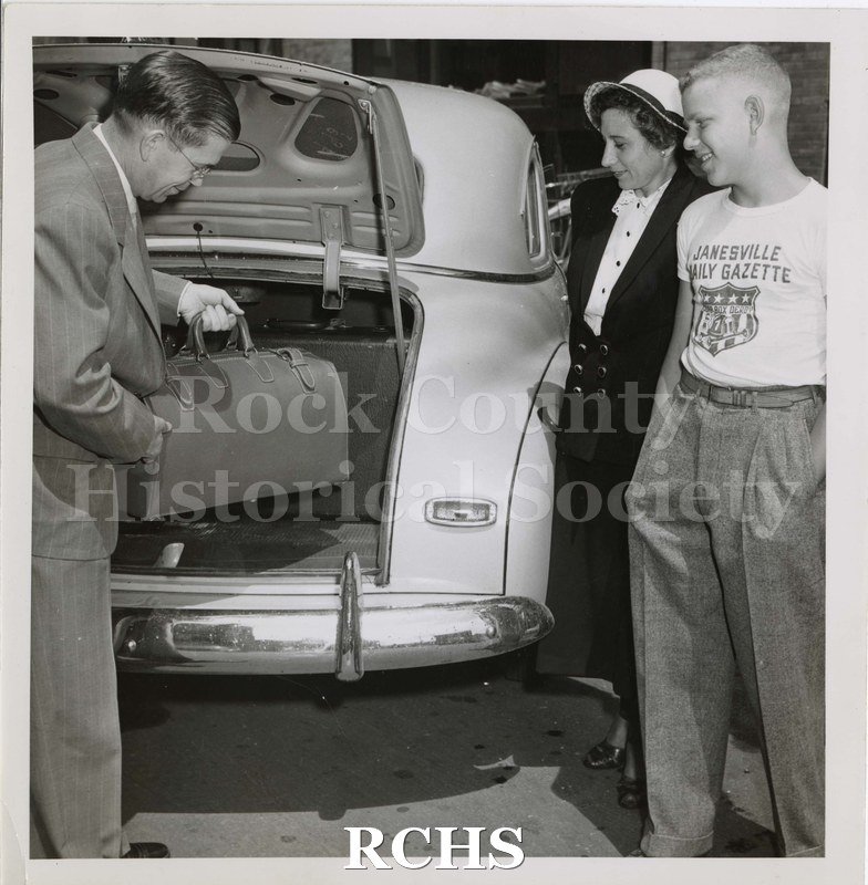 Black and white photograph of Mr. Ralph O. Hausner putting a bag in the trunk of his car while Carliss B. Shuler and Mrs. Ralph O. Hausner watch.