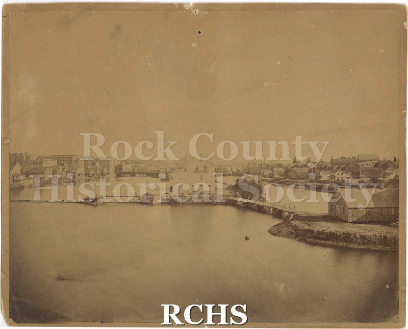 Tinted photo of Rock River in downtown Janesville. Mill run and Fords Mills visible; wagon and horse in middle left.