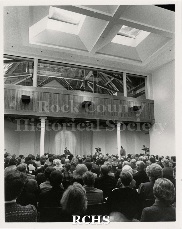 Black and white photograph of Pearson's Hall rededication. Taken on the ground floor. A crowd of people sit and listen to the speaker. Cameras at the front.