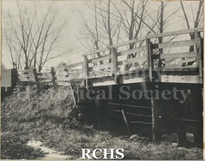 Black and white photograph of the Jackson Street Bridge between late 1800's 1930. Shows bridge with wooden railings; below is barbed wire attached to poles.