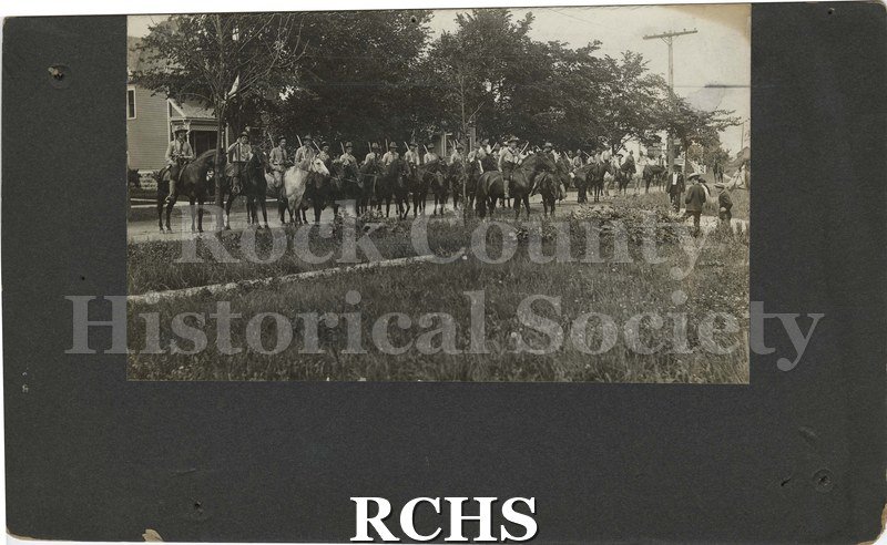 Black and white photograph of a line of men mounted on horses holding sabers. House visible in the background and a sign reading Clarence (?) Street.