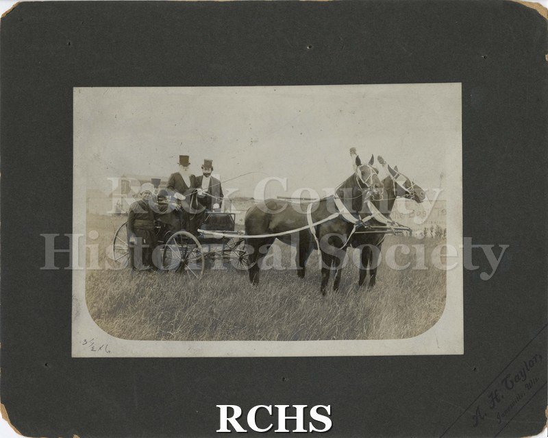 Black and white photograph of two men in a carriage drawn by two horses. They wear suits and top hats. One carries a whip.