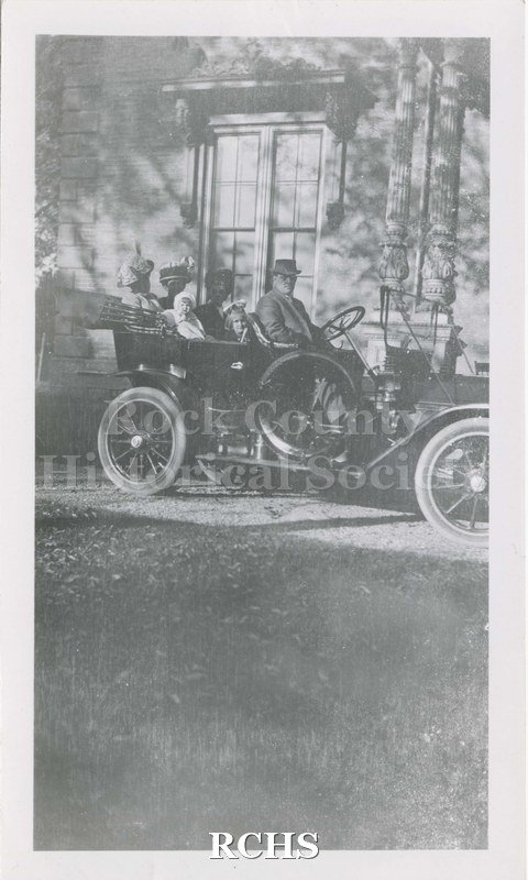 Black and white photograph of family members seated in 1920s era automobile. Shows four adults and two children in an old automobile parked in front of home(?).