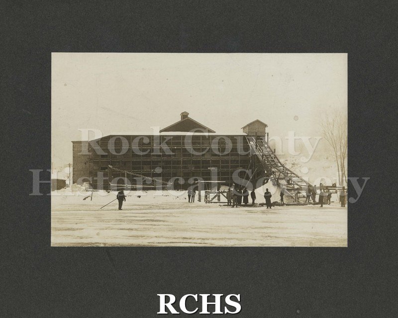 Black and white photograph of the Ice Harvest in Janesville, WI ca 1907. There are several people standing on the ice watching the ice cutting.