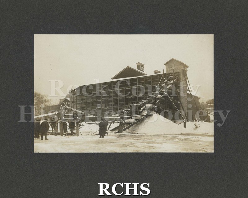 Black and white photograph of the Ice Harvest in Janesville, WI ca 1907. There are several people standing on the ice watching the ice blocks go up the conveyor.
