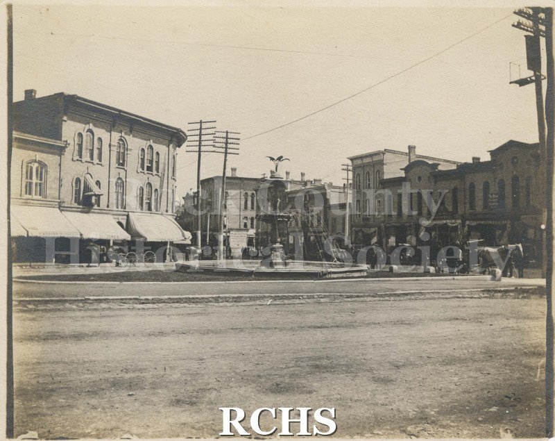 Black and white photograph of the Corn Exchange. Milwaukee & Fanklin Sts. Shows statute in middle of polygon with buildings to the left and right.