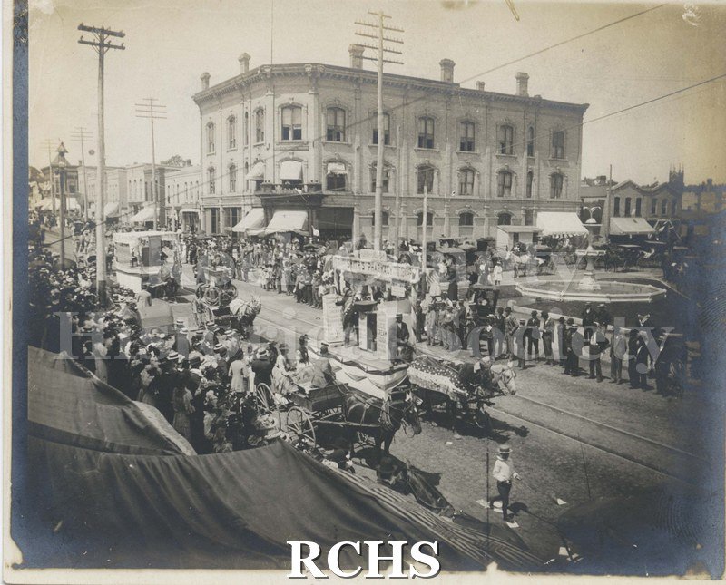 Black and white photograph of the Parade of Floats on West Milwaukee Ave. Parade of floats is heading east, passing what is likely the fountain at the Corn Exchange.