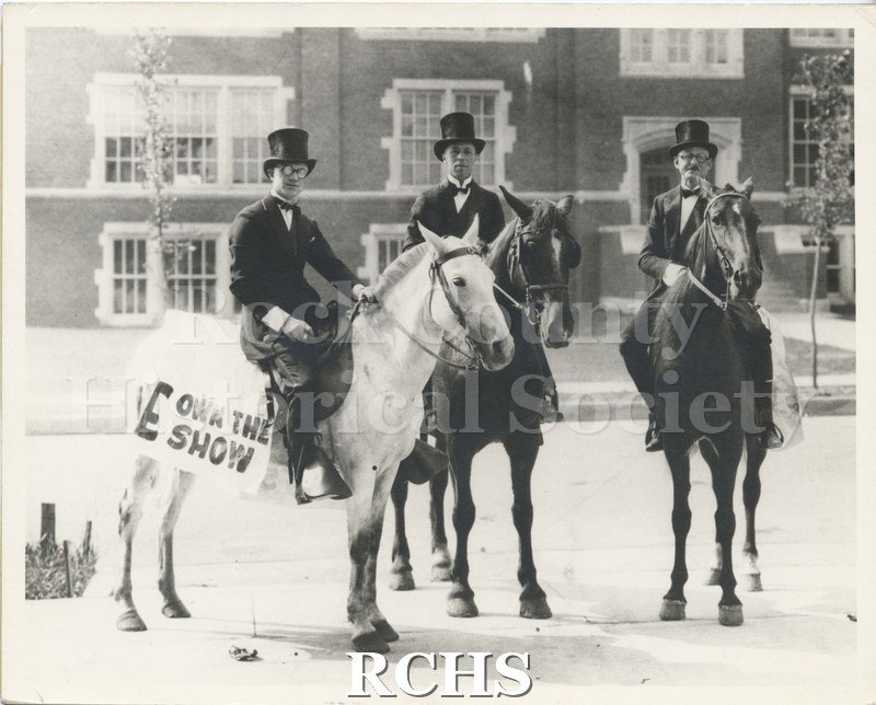 Black and white photograph of three horsemen. Shows three men on horseback wearing top hats. In background, possibly Janesville High School.