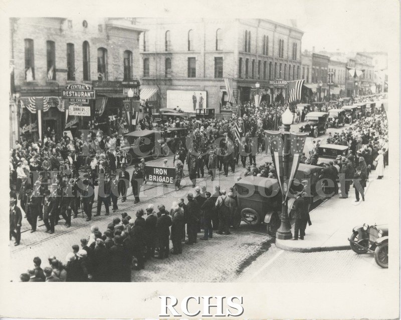 Black and white photograph of Iron Brigade Parade, likely on W. Milwaukee St. Street, Janesville. Onlookers and period automobiles line the street.