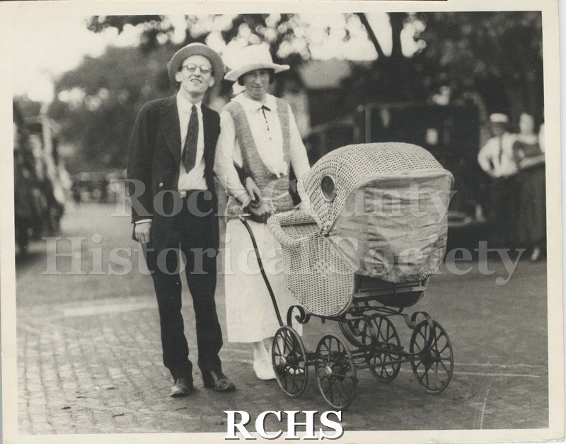 Black and white photograph of Nonesuch Circus Parade. Shows a man and woman pushing a wicker baby carriage along brick-paved street.
