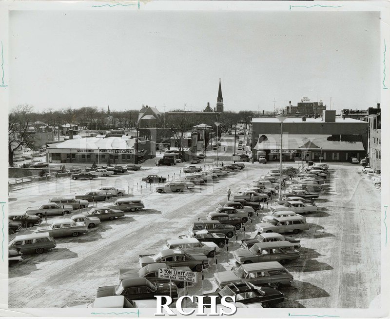 Black and white photograph of Janesville Parking Plaza over Rock River, looking west. Shows Adams Furniture Co.