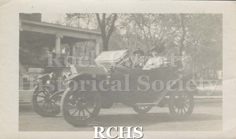 Black and white photograph of Mrs. Jeffris and Kenneth Jeffris. Shows a man and woman in vintage automobile parked on residential street. Written on reverse: "Mrs.