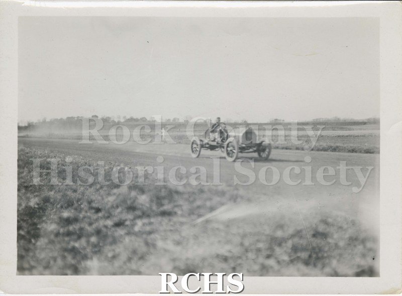 Black and white photograph of a man driving race car. Written on reverse: "Practice run at Fair grounds 1911".