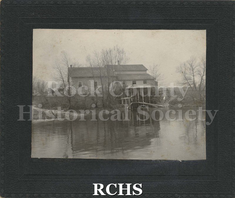 Black and white photograph of Fulton, WI. Shows the back of a building with pilings into the river. On left side are two horses on higher ground.