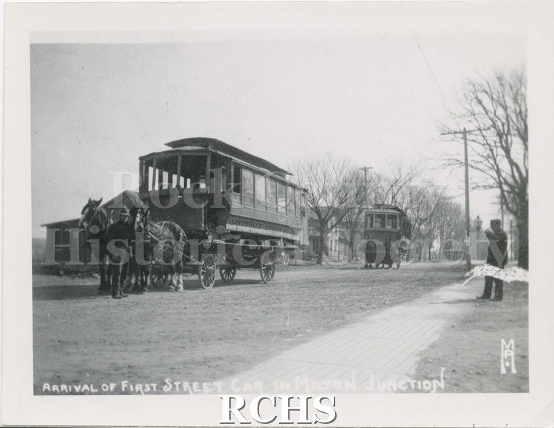 Black and white photograph of the "Arrival of First Street Car in Milton Junction". Shows two street cars mounted on two wagons pulled by a team of horses.