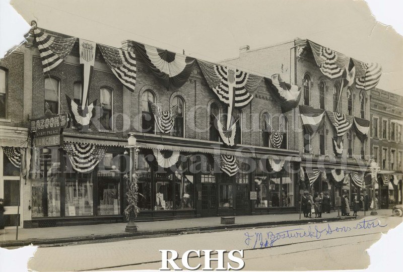 Black and white photograph of the storefronts on Main Street in Janesville, WI. Shows bunting displayed on buildings.