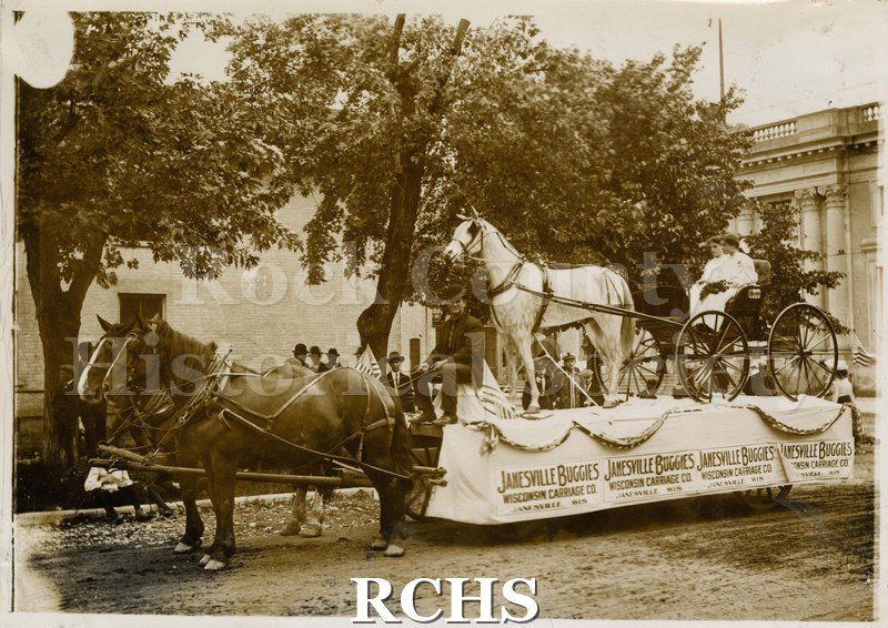 Black and white photograph of a horse-drawn parade float sponsored by Janesville Buggies Wisconsin Carriage Co. Carnegie Library visible in background.