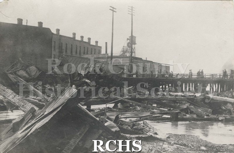 Black and white photograph of the Janesville, WI, fire April 1913. Shows buildings in background with river in front.