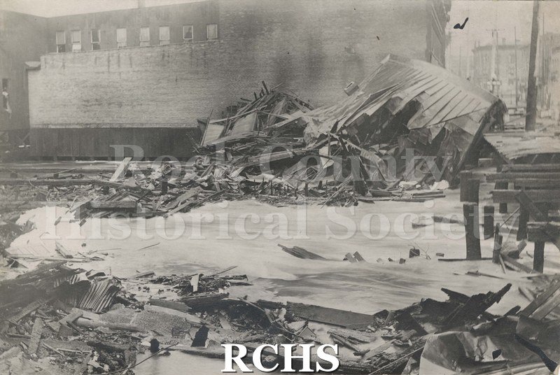 Black and white photograph of the Janesville, WI, fire April 1913. A building that has collapsed is shown in the middle of the picture with debris in front.