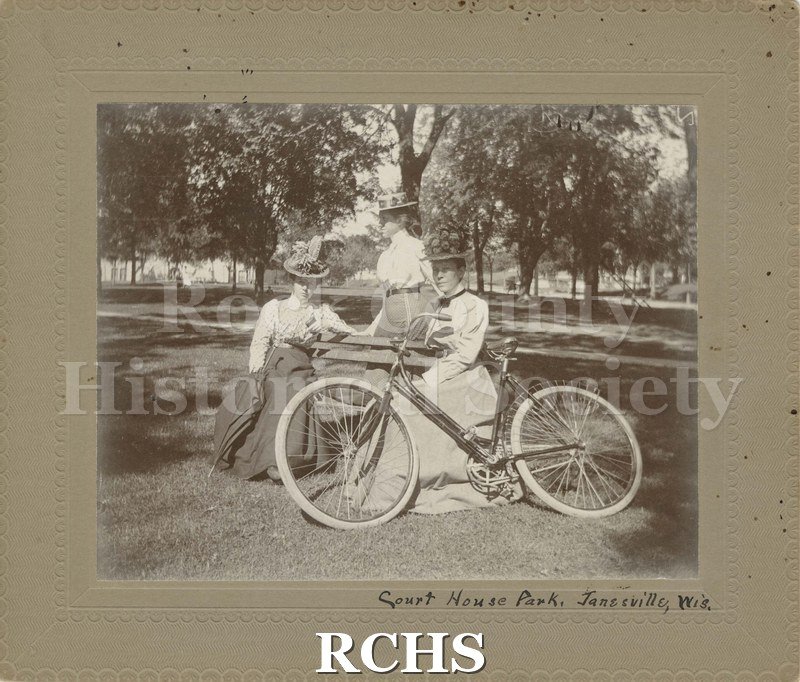 Black and white photograph of three women in hats seated on a park bench with bicycle beside them. Two other figures in distance.